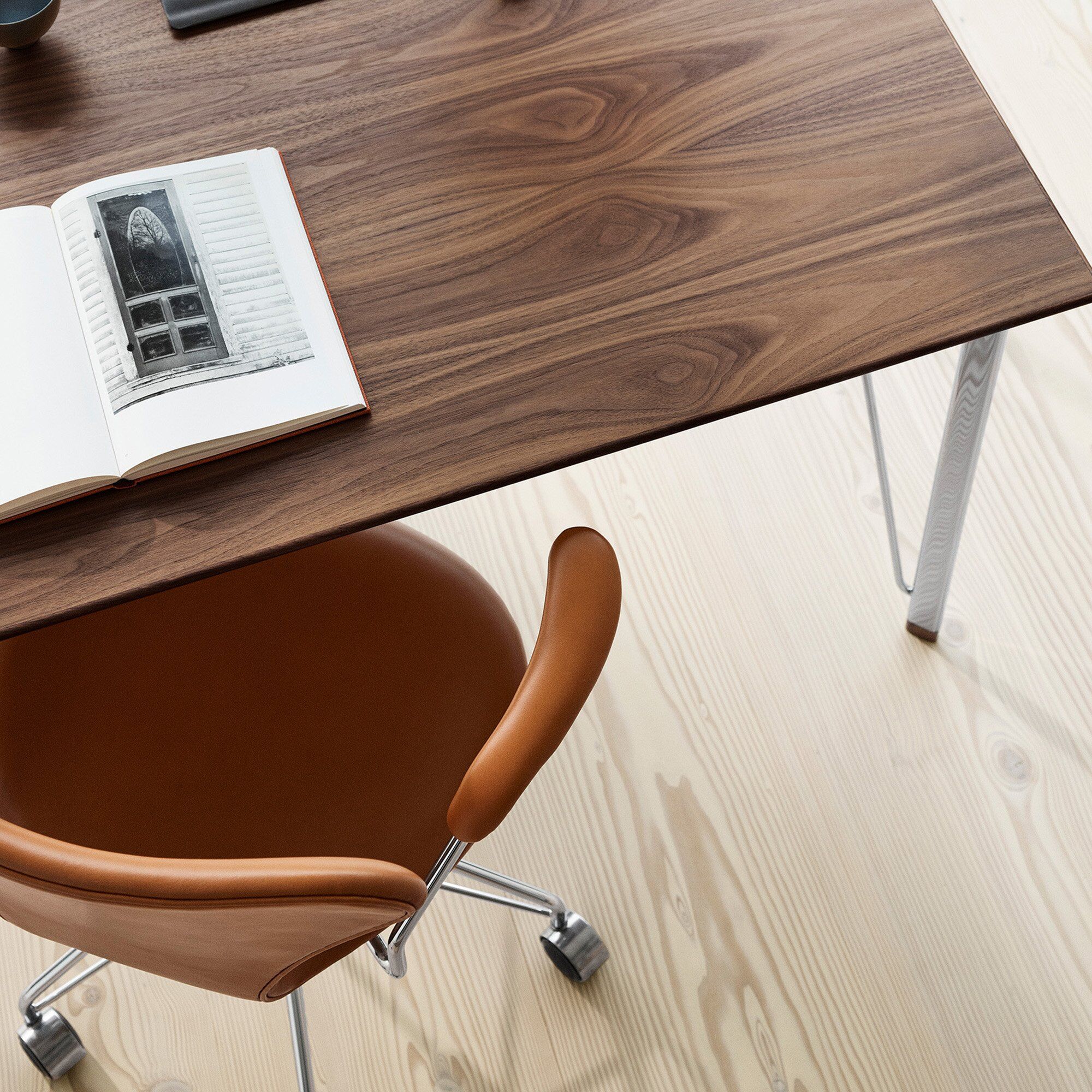 Detailreiches Foto eines modernen, rotbraunen Bürostuhls »Series 7™« von Fritz Hansen mit Lederbezug und Armlehnen an einem Schreibtisch aus dunklem Holz. Auf dem Tisch liegt ein aufgeschlagenes Buch mit einer Schwarzweißaufnahme. Der Boden ist helles Holz.