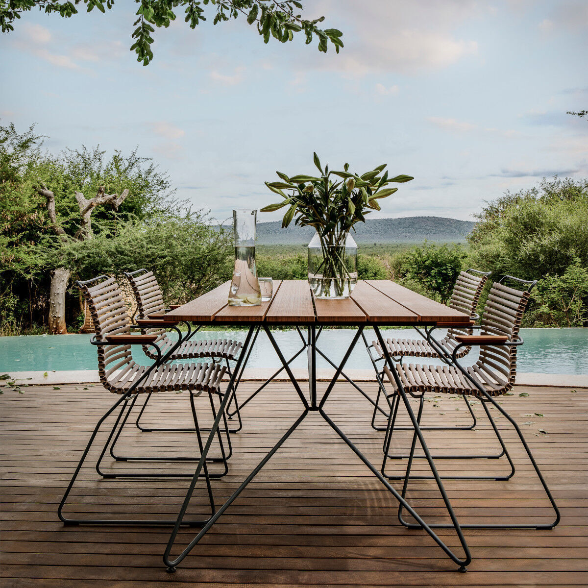 Ein moderner Gartentisch »Sketch« steht auf einer Terrasse mit Holzboden. Rund um den Tisch stehen Stühle mit Metallrahmen und Holzlatten. In der Mitte des Tisches befindet sich eine Blumenvase mit Blumen. Im Hintergrund sieht man einen Pool und grüne Natur.