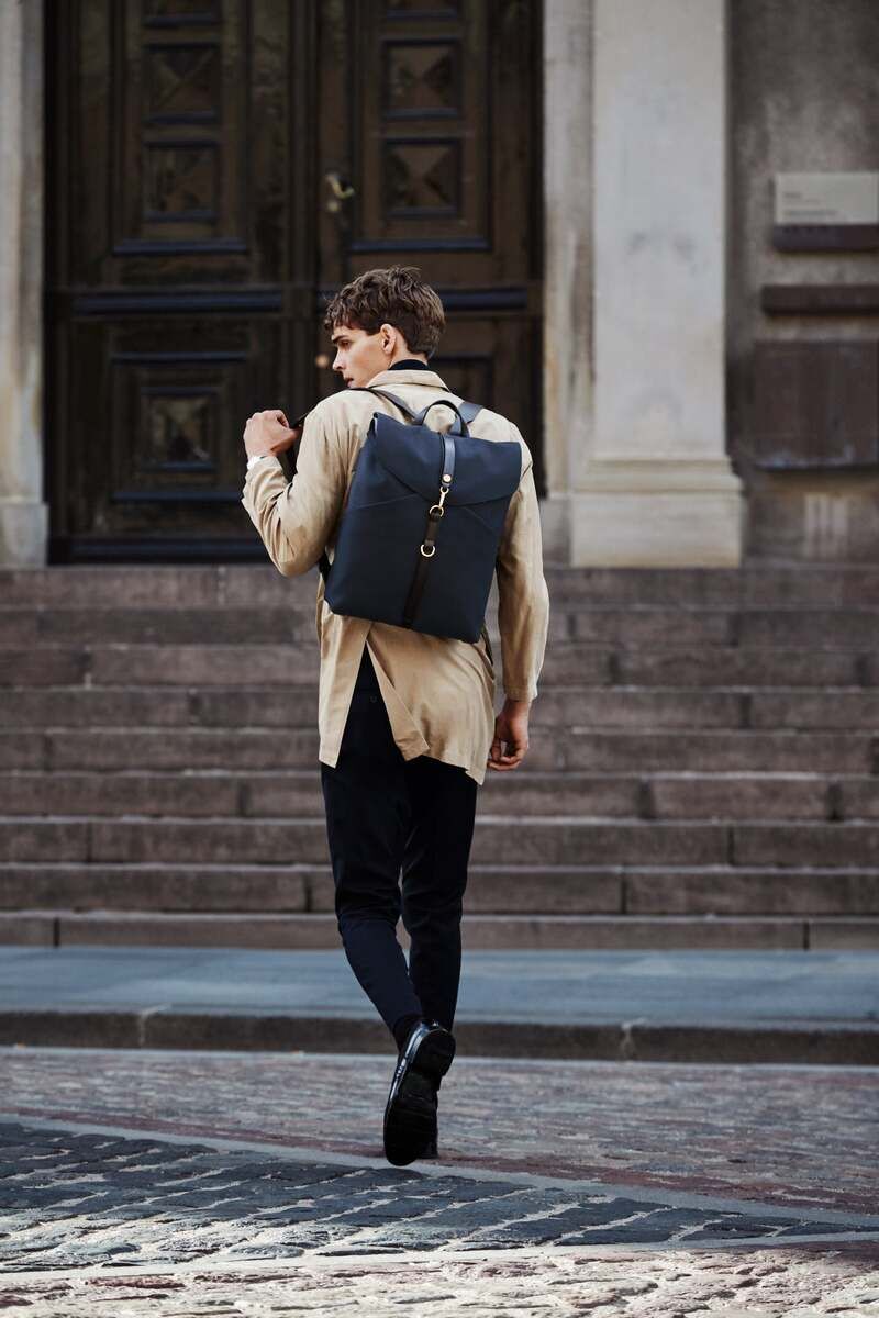 Ein Mann trägt einen hochwertigen, schwarzen Rucksack von MISMO. Er geht eine steinerne Treppe mit einer stilvollen Tür im Hintergrund hinauf. Der Rucksack hat ein minimalistisches Design und passt gut zu seiner beigen Jacke und schwarzen Hose. Die Szene spielt sich in einer städtischen Umgebung ab.
