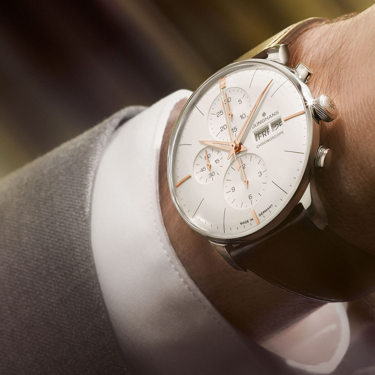 Eine stilvolle Junghans ZEIT-Sonderedition Meister Chronoscope Herrenuhr mit einem weißen Zifferblatt, silbernem Edelstahlgehäuse und braunem Lederarmband wird am Handgelenk getragen; im Hintergrund unscharf.