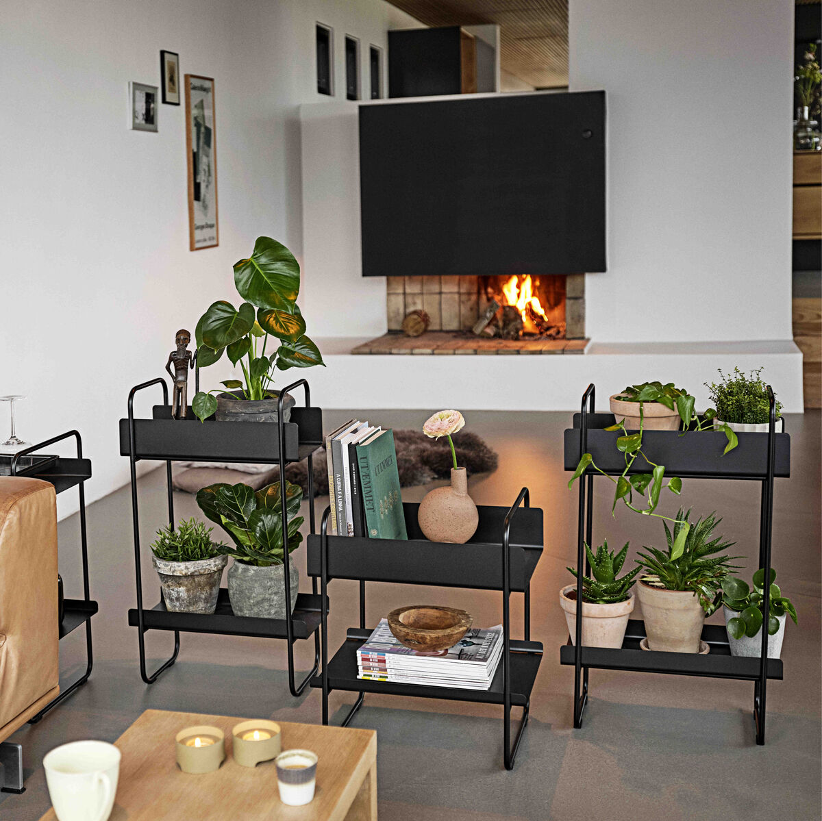 Ein modernes Wohnzimmer mit einem Leder-Sofa, mehreren schwarzen Konsolen- und Pflanztischen mit grünen Pflanzen, Büchern und Kerzenhalter vor einem Kamin.