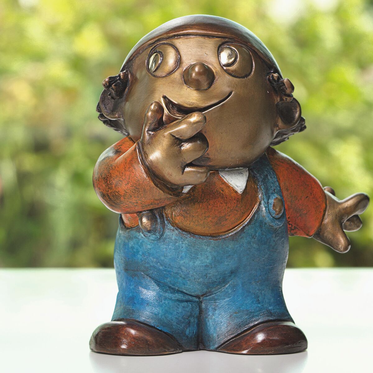 Sehr detailreiche Bronze-Skulptur eines Mainzelmännchens mit blauer Latzhose, orangener Jacke und weißem Hemd. Die Figur steht auf einer hellen Fläche vor grünem, unscharfem Hintergrund. Mimik und Oberfläche sind sorgfältig ausgearbeitet und handpatiniert.