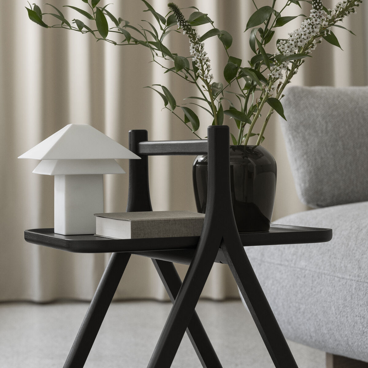Der schwarze Beistelltisch "ETA" von blomus steht vor einem beigefarbenen Vorhang und neben einem grauen Sofa. Auf dem Tisch befinden sich eine weiße Designlampe, ein Buch sowie eine schwarze Vase mit grünen Zweigen und weißen Blüten.