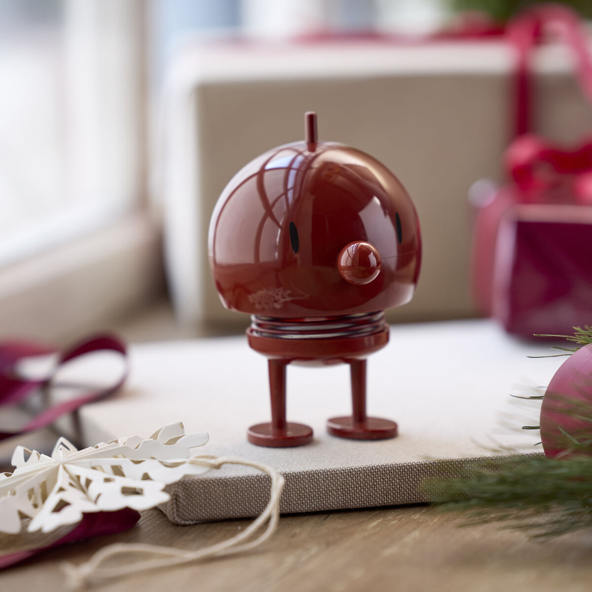 Der Hoptimist Bumble Maroon ist mittig auf einem Tisch aus Holz vor Fensterscheibe platziert. Im Hintergrund liegen bordeauxrote Geschenkboxen mit weißen Schleifen. Daneben Deko wie Tannenzweig, Kugel, weiße Schneeflockenanhänger und ein Band.
