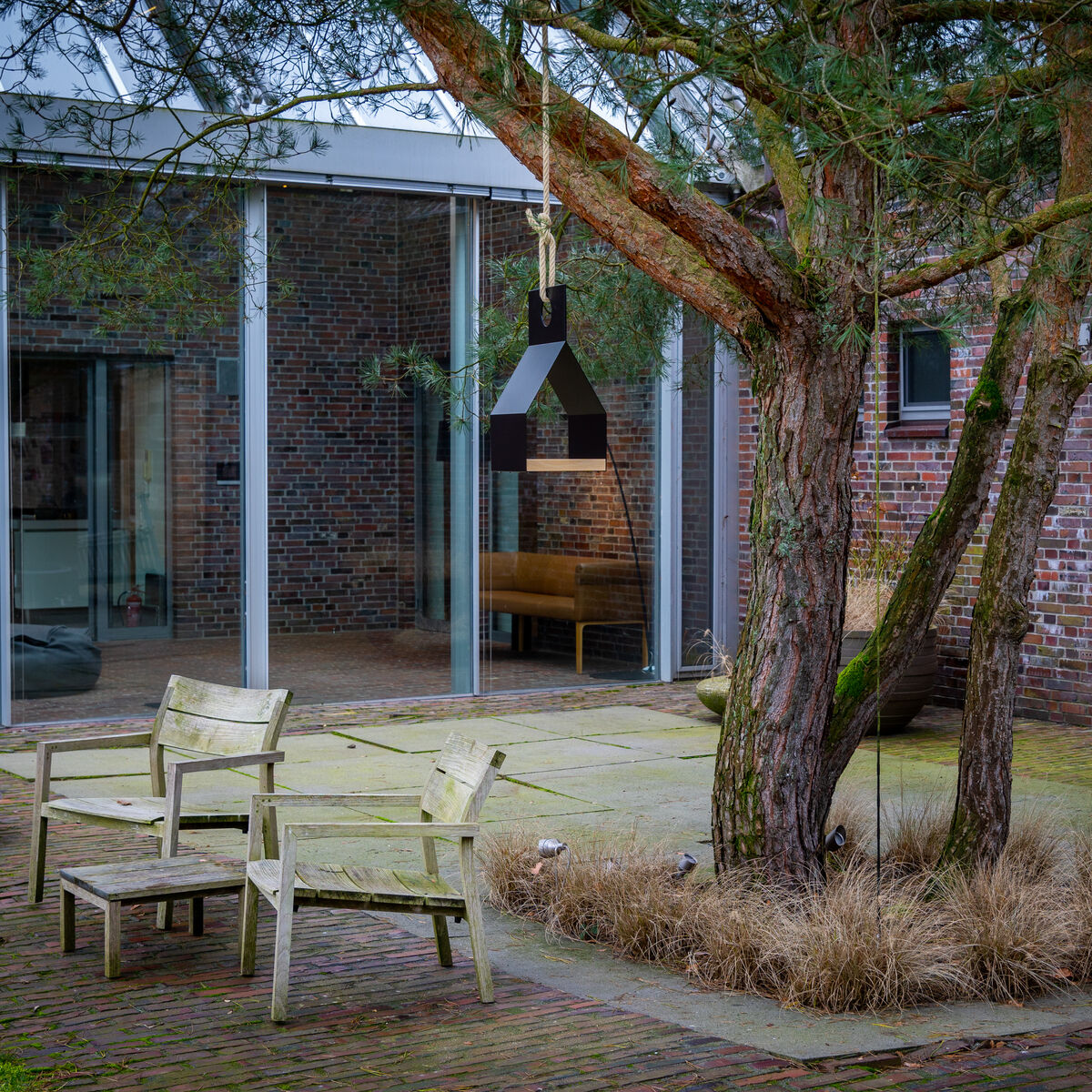 Im Bild ist ein moderner Hinterhof mit gepflasterter Terrasse und Sitzgruppe aus hellem Holz zu sehen. Zwischen zwei Baumstämmen hängt das Vogelhaus »FLY« der Marke finbeaux. Die Umgebung besteht aus Rasen, Ziergräsern und Backsteinmauern mit großen Glasfenstern.