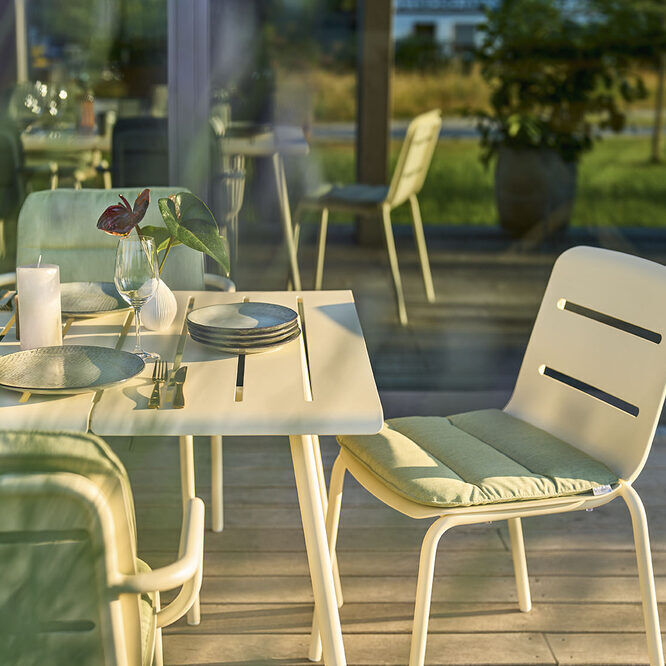 Ein Esszimmer- oder Gartenmöbel-Stuhl namens VAPIO ist in einem modernen, minimalistisch eingerichteten Essbereich auf einer Terrasse zu sehen. Der Stuhl ist weiß mit grünen Kissen, platziert an einem gedeckten Tisch mit Kerzen und Geschirr.