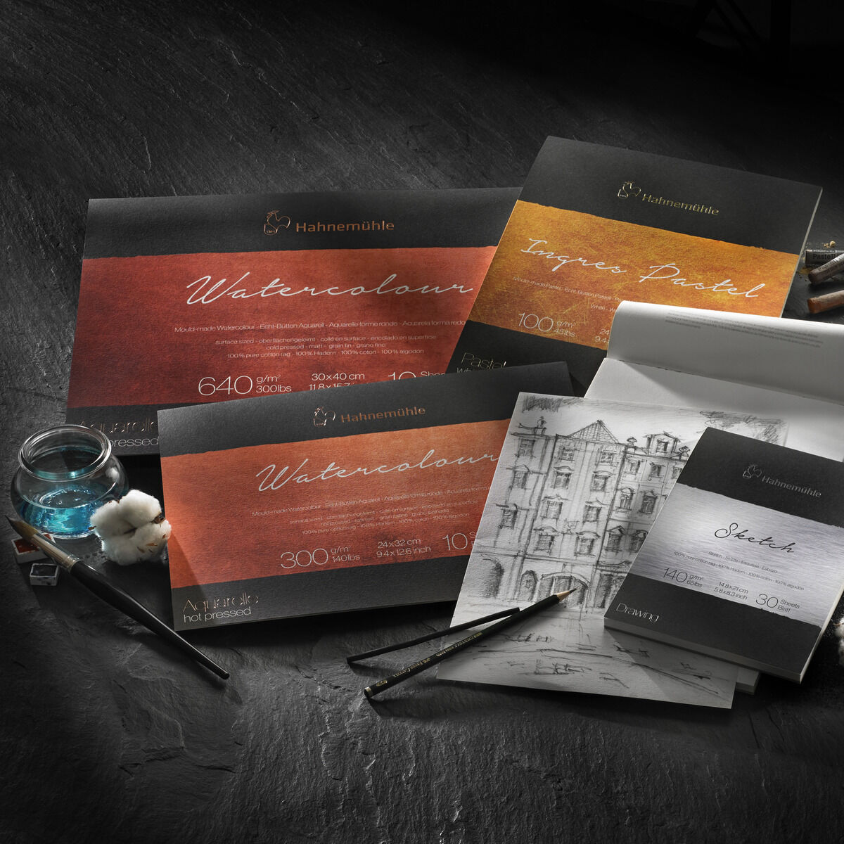 Ein Arrangement von Hahnemühle Aquarellpapier-Produkten, darunter ein Aquarellblock und Skizzenblock. Die Verpackungen sind schwarz mit roten, orangefarbenen und braunen Etiketten, umgeben von Kunstutensilien wie Pinsel und Tinte auf einem dunklen Hintergrund.