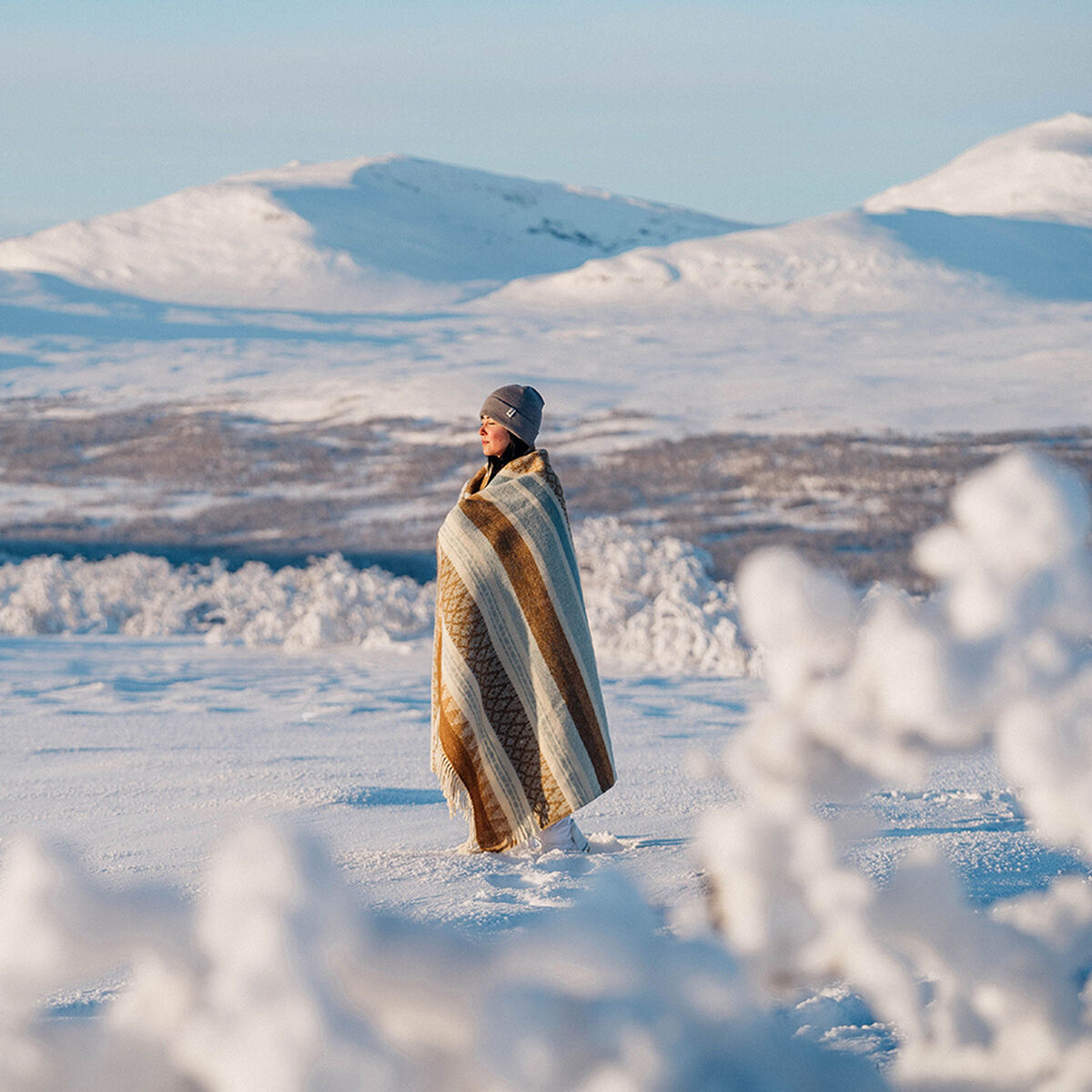 Eine in eine Decke »Freja« gehüllte Person steht in einer verschneiten Berglandschaft mit weißem Schnee und entfernt liegenden Bergen unter einem klaren, blauen Himmel.