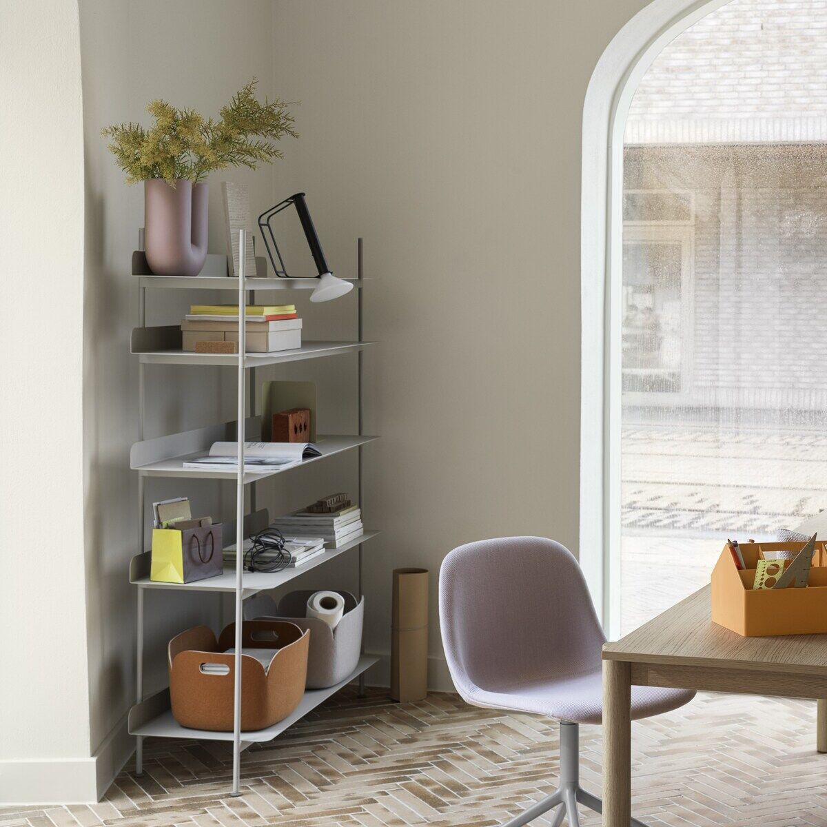 Ein modernes und minimalistisches Home Office mit einem großen Fenster, das viel Tageslicht hereinlässt. Ein Schreibtisch mit diversen Büromaterialien und eine Tragbare Lampe »Piton« auf der Oberfläche. Eine Regalwand mit mehreren Gegenständen und Dekorationen steht im Hintergrund.