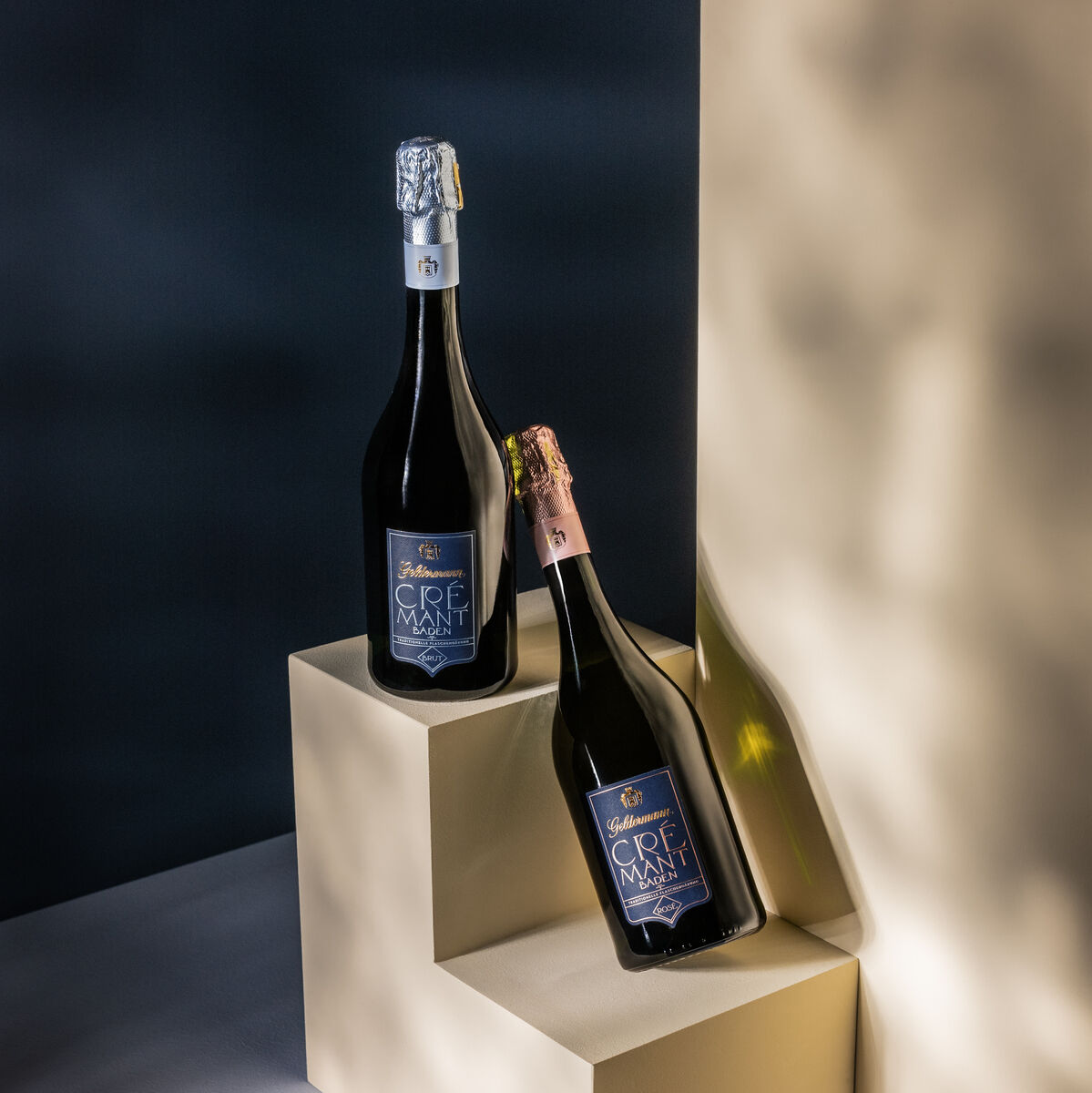 Zwei Flaschen Crémant mit Neoprenmanschetten auf beigen Stufen vor einem dunkelblauen Hintergrund platziert.