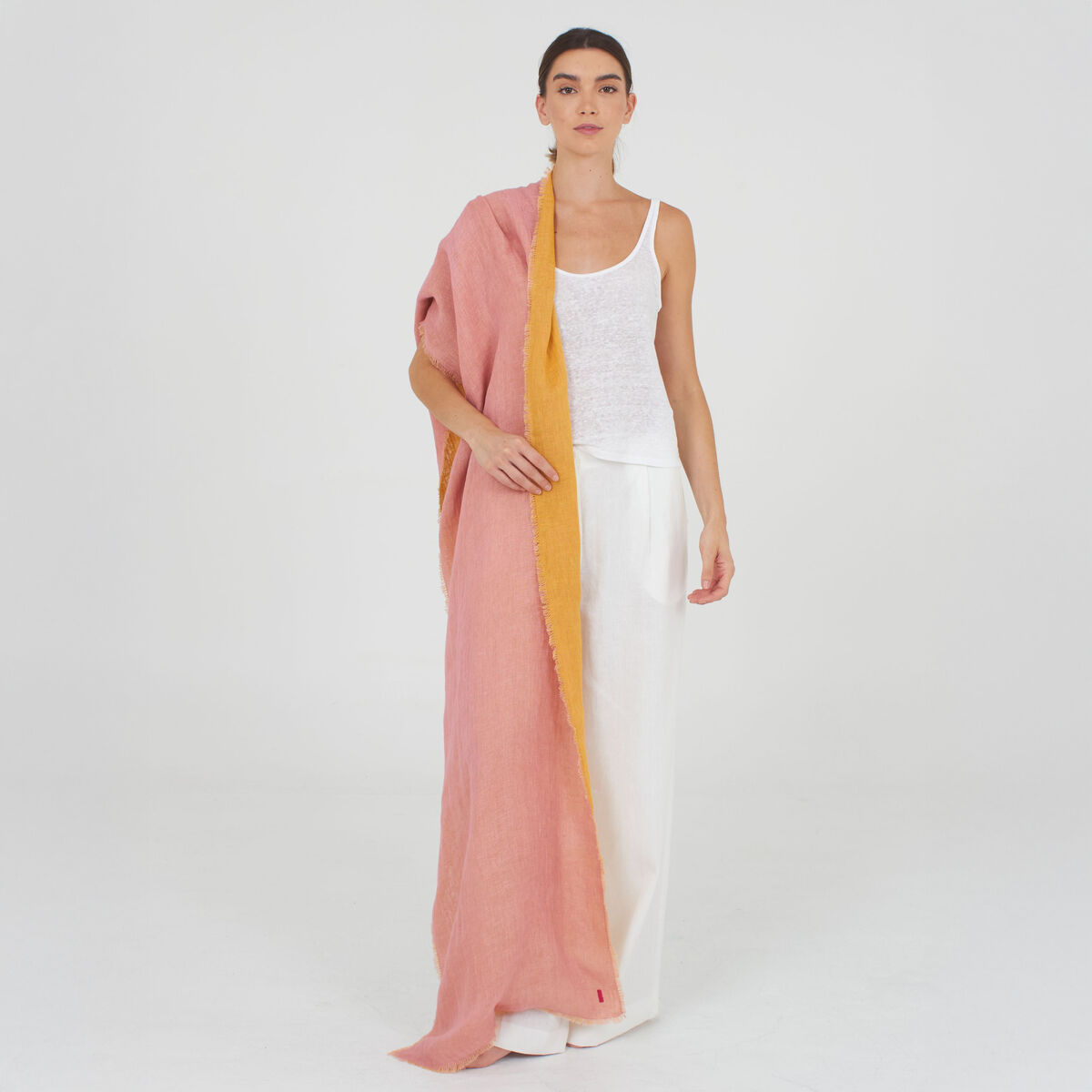 Eine Frau trägt einen großzügigen Leinenschal »Alsi« von Shalls Barcelona, Farbe Pink mit gelbem Rand, locker um die Schultern gelegt. Sie trägt ein weißes Oberteil und eine weite weiße Hose. Der Schal wirkt leicht, weich und modisch in einem minimalistischen, hellen Studio.