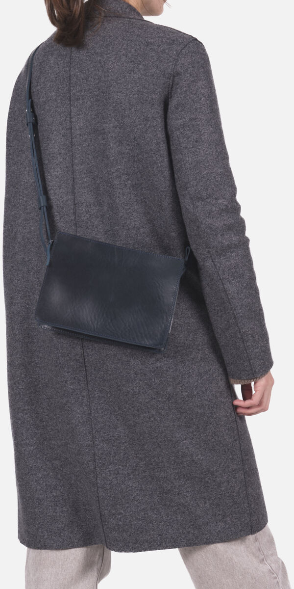 Frau mit dunkelblauer Kernleder-Umhängetasche, getragen über der Schulter, bekleidet mit grauem Mantel und hellen Hosen vor hellem Hintergrund. Die Tasche ist schlicht, rechteckig und passt zu modernen, minimalistischen Outfits.