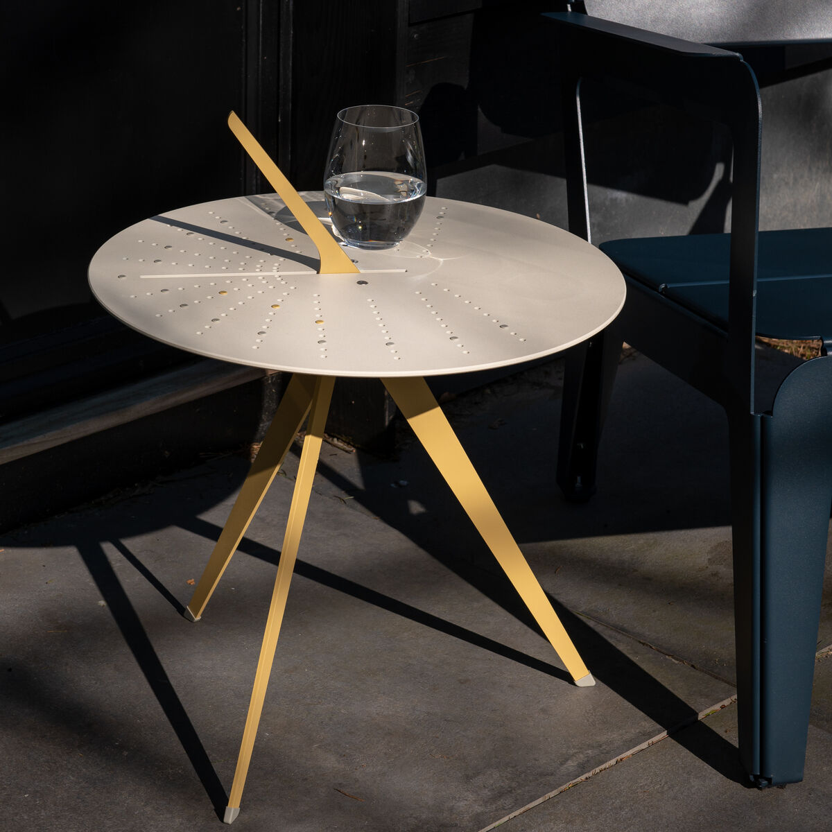 Moderner Tisch »Sundial« mit runder, beiger Tischplatte und gelben Beinen, draußen neben einem dunkelgrauen Stuhl auf einer Steinfläche. Ein Glas Wasser steht darauf im Sonnenlicht.