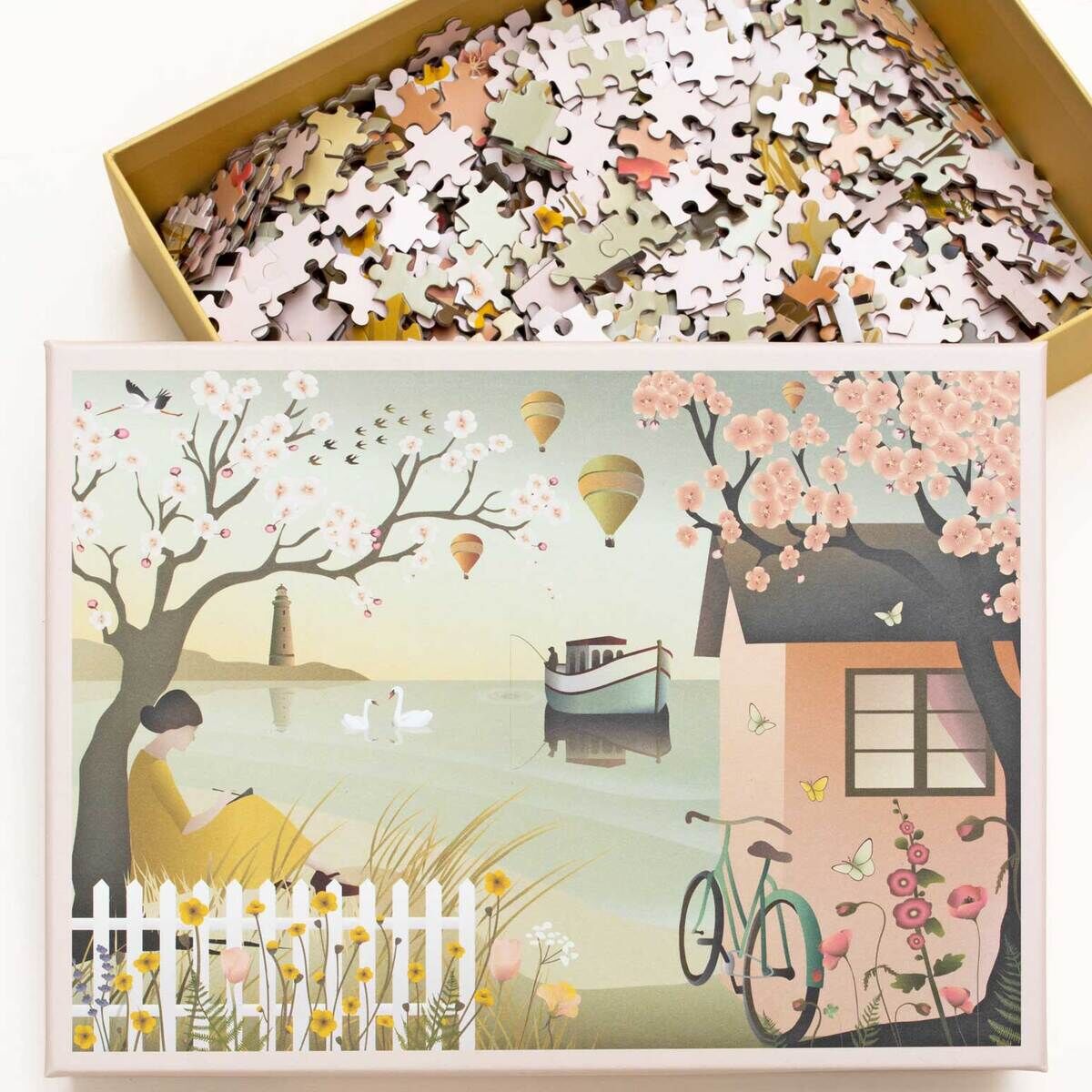Ein Puzzle mit dem Motiv 'By the sea', das eine ruhige Landschaft zeigt. Zu sehen sind ein kleines Haus mit blühenden Bäumen, ein Fahrrad, ein Boot auf dem Wasser und Heißluftballons am Himmel. Im Vordergrund sitzt eine Person am Ufer. Die Farben sind sanft und pastellig.