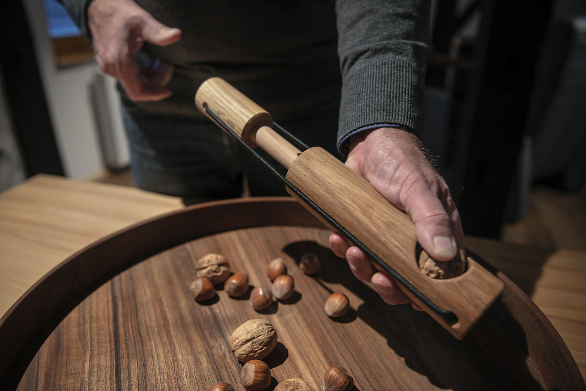Ein hölzerner Nussknacker namens »Kracher« wird von einer Person benutzt, um Walnüsse zu knacken. Der Nussknacker liegt auf einer Holzschale zusammen mit Walnüssen und Haselnüssen; eine männliche Hand hält das Gerät. Verschiedene Brauntöne dominieren das Bild.