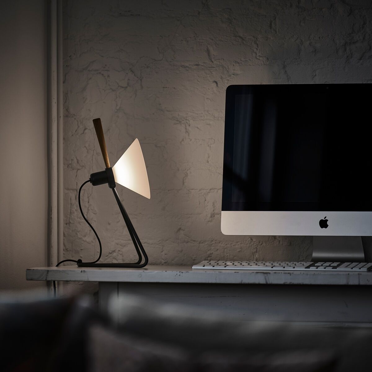 Eine Tisch-/Hängelampe namens »Spot«, die auf einem Schreibtisch neben einem Computer steht. Die Lampe hat ein modernes Design mit einem schwarzen Ständer und einem weißen Schirm. Die Wand im Hintergrund ist in einem neutralen Grauton gehalten.