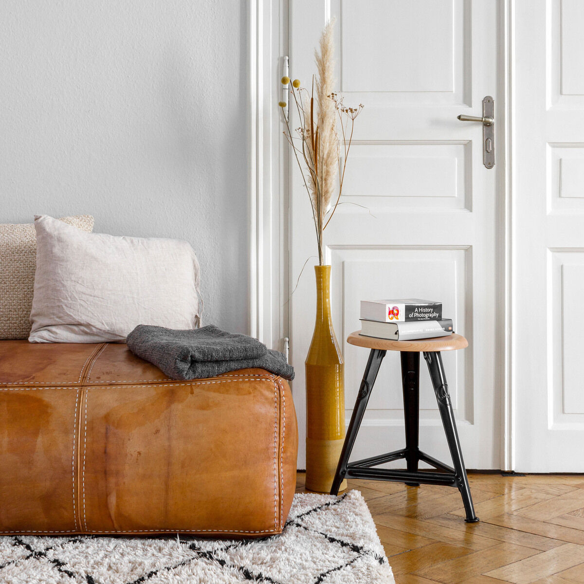 Wohnzimmer mit einem Ledersofa, dekoriert mit Kissen; daneben steht ein kleiner Schemel aus Eiche mit mattschwarzem Rahmen und darauf liegt ein Buch. Im Hintergrund steht eine weiße Tür und eine Vase mit getrockneten Pflanzen. Der Raum hat einen holzfarbenen Parkettboden und einen hellgemusterten Teppich.