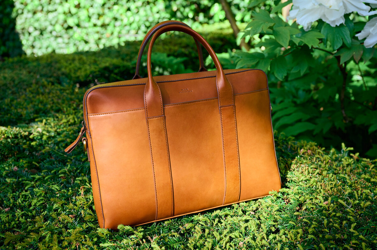 Eine braune Ledertasche »Harvey« steht auf grüner Vegetation, umgeben von Pflanzen im Hintergrund. Die Tasche hat zwei robuste Griffe und ein elegantes Design.
