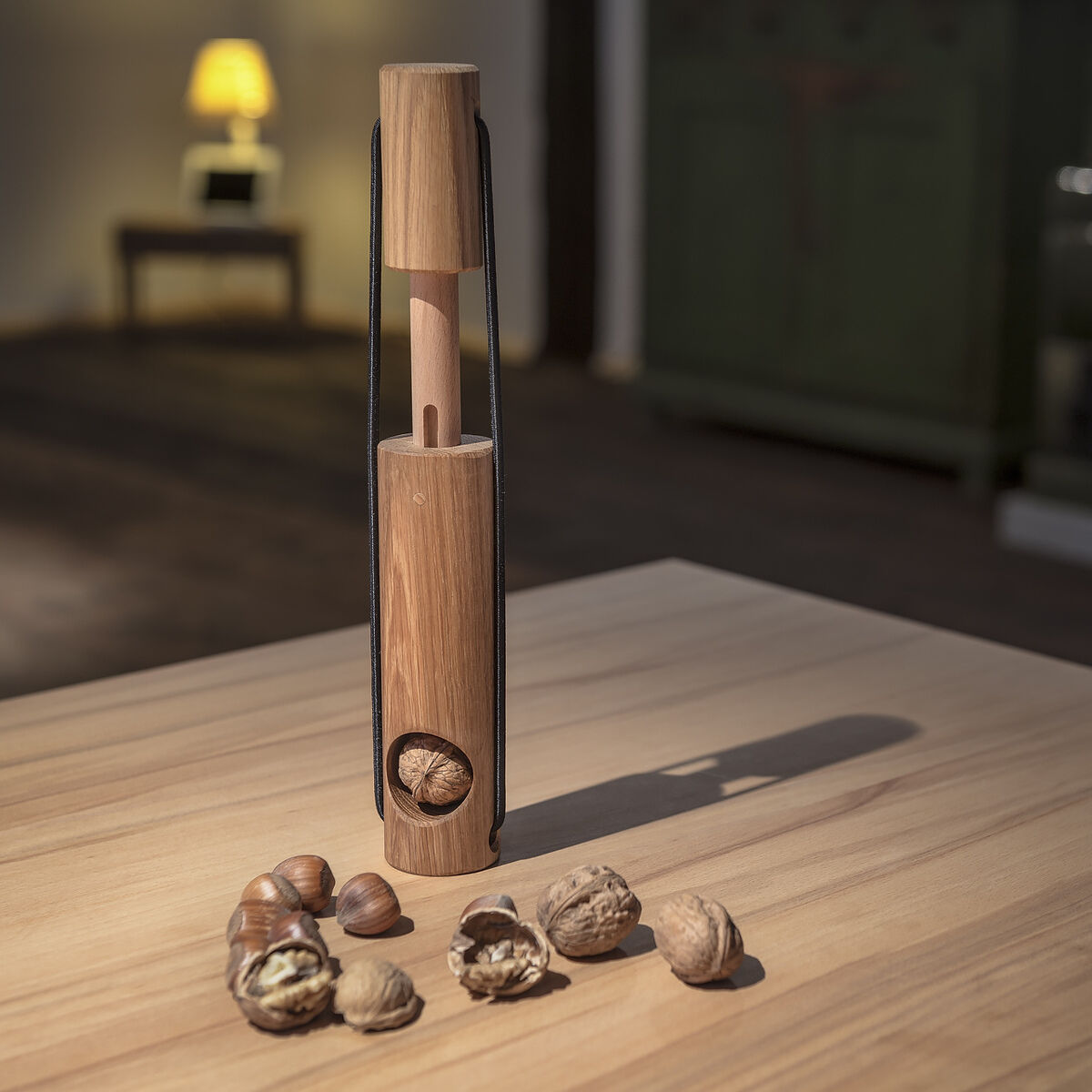 Ein eleganter Nussknacker aus Holz mit einem ergonomischen Design steht auf einem Holztisch. Um den Nussknacker herum liegen geöffnete und ganze Walnüsse. Im Hintergrund sind unscharfe Details eines Raumes mit einem Sofa und einer Lampe sichtbar.