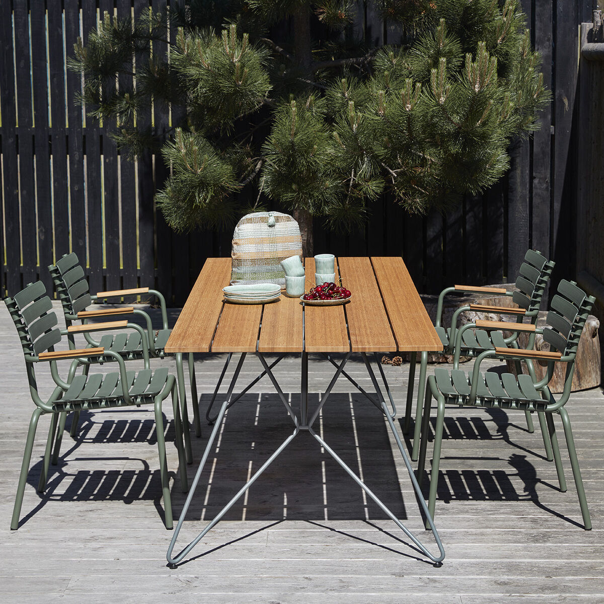 Terrassenmöbel-Set mit einem Holztisch und sechs grünen Metallstühlen mit Holzarmlehnen, auf einer Holzterrasse mit einem Baum im Hintergrund.