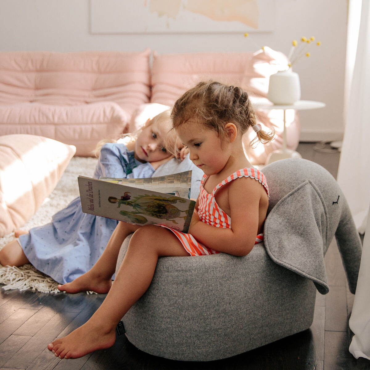 Zwei kleine Mädchen sitzen in einem gemütlichen Zimmer. Eines der Mädchen sitzt auf einem grauen Rollelefant und liest ein Buch. Das andere Mädchen lehnt sich an einen rosa Sofa und schaut zu. Im Hintergrund sind eine Stehlampe, eine Decke und ein Gemälde zu sehen.