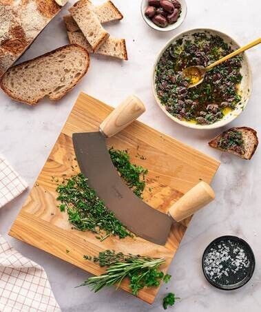 Wiegemesserbrett 'Soul' aus Holz mit Kräutern und einem Wiegemesser, umgeben von Brot, Oliven und einer Schüssel mit Kräutermischung auf einer Marmoroberfläche.