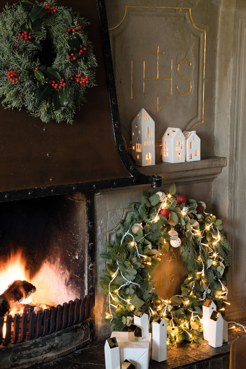 Ein festlich dekorierter Kamin mit brennendem Feuer. Links und rechts sind große grüne und rote Kränze. Auf dem Kamin Sims sind weiße Lichthäuser angeordnet, die warmes Licht ausstrahlen.