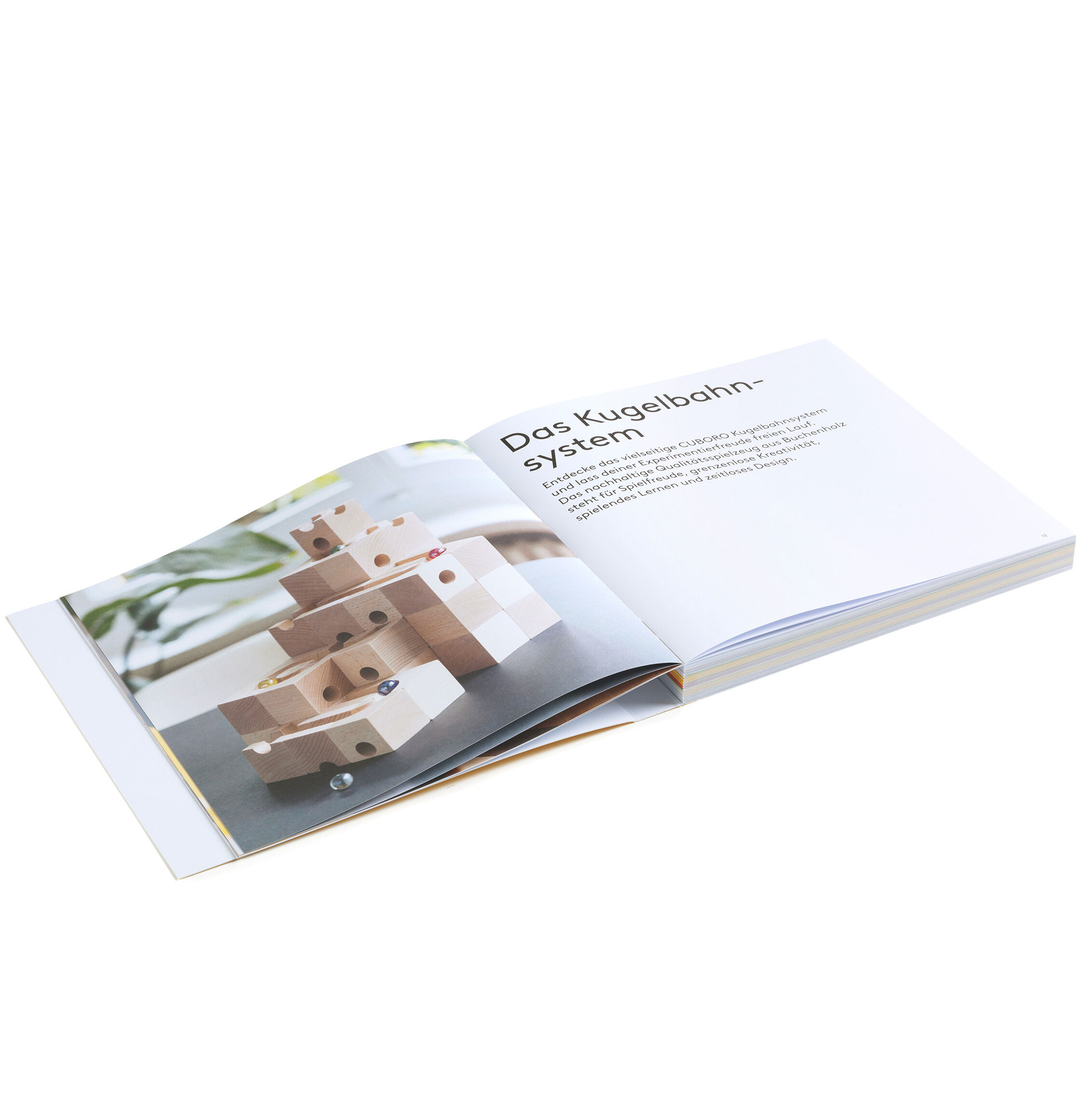 Aufgeschlagenes Buch mit einer Bauanleitung für die Cuboro-Kugelbahn. Links befindet sich ein großes Foto eines aus Holzbausteinen gebauten Kugelbahnsystems, rechts steht der Titel "Das Kugelbahnsystem" und eine kurze Erläuterung in deutscher Sprache. Heller, klarer Hintergrund.