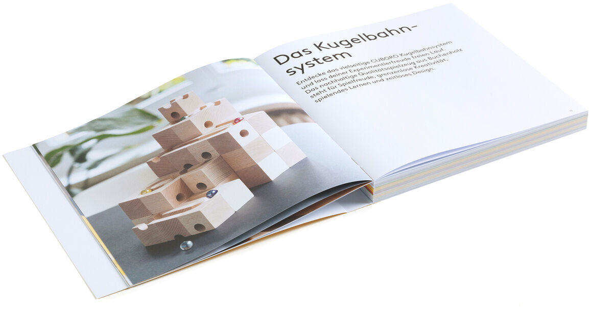 Aufgeschlagenes Buch mit einer Bauanleitung für die Cuboro-Kugelbahn. Links befindet sich ein großes Foto eines aus Holzbausteinen gebauten Kugelbahnsystems, rechts steht der Titel "Das Kugelbahnsystem" und eine kurze Erläuterung in deutscher Sprache. Heller, klarer Hintergrund.