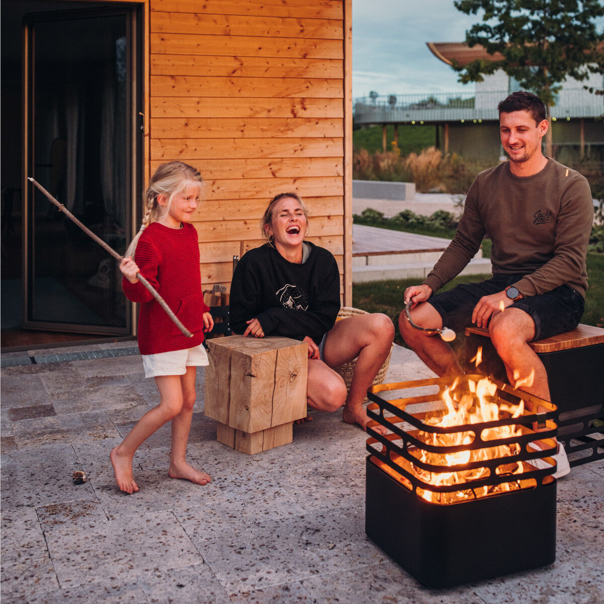 Ein Mann, eine Frau und ein Mädchen sitzen im Freien neben einer brennenden Feuerschale. Sie lachen und genießen den Abend. Im Hintergrund sieht man ein Holzgebäude und eine Terrasse.