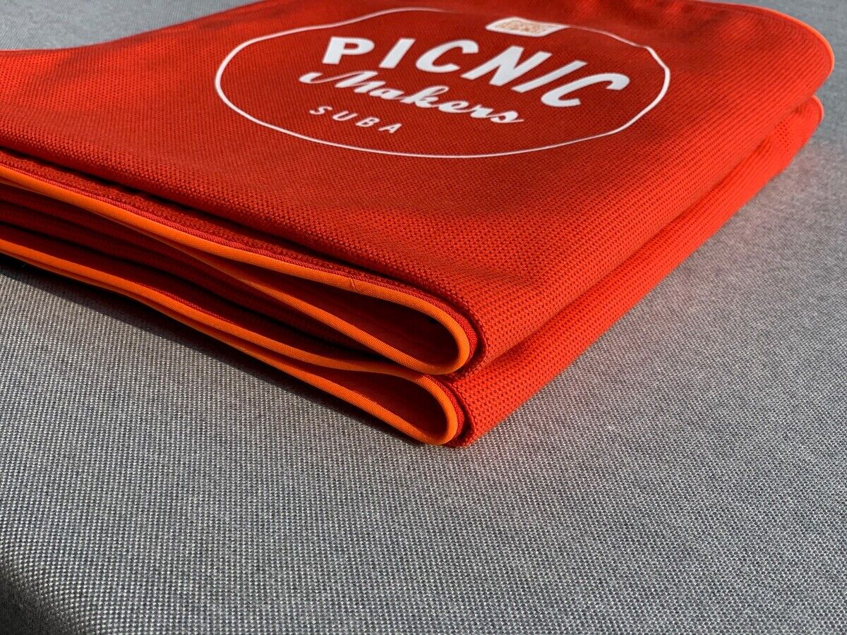 Eine gefaltete Picknickdecke in leuchtendem Rot mit der Aufschrift 'PICNIC Makers SUBA' liegt auf einer grauen Oberfläche.
