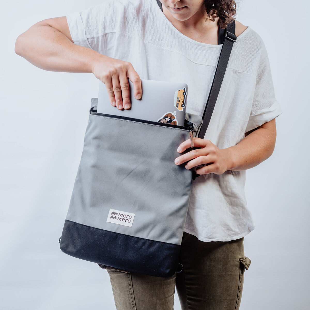 Eine Person hält eine graue und schwarze Stofftasche namens »Meije«, die ein Tablet und ein kleines Gerät enthält. Die Person trägt ein weißes T-Shirt und eine olivgrüne Hose.