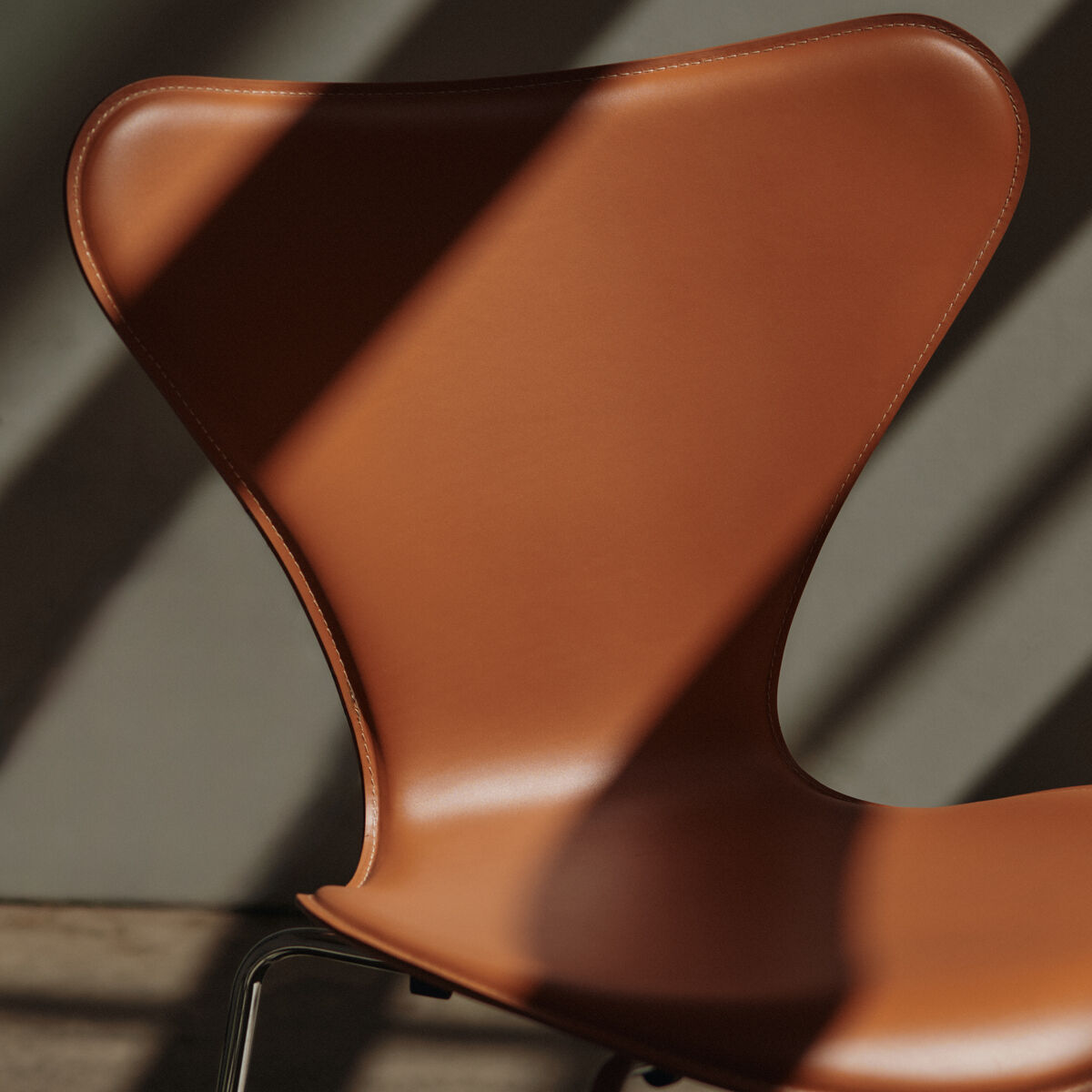 Detaillierte Nahaufnahme eines modernen Designerstuhls, Modell Series 7™ Tailored von Fritz Hansen, mit sattelbraunem Lederbezug und glänzenden Chrombeinen. Der Stuhl steht vor einer neutralen Wand, durch die Streiflicht Schatten auf die Rückenlehne wirft.