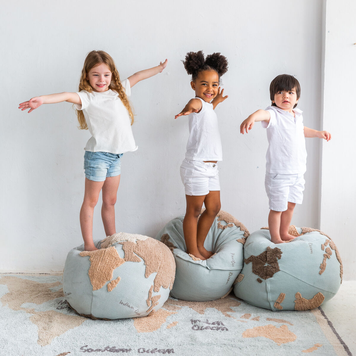 Drei Kinder stehen auf Sitzkissen mit Weltkartendesign, lächelnd und die Arme ausbreitend vor einer weißen Wand.