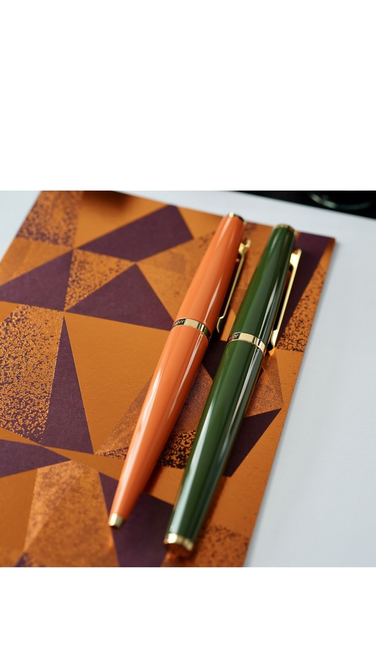 Kugelschreiber »Design06« Orange und Grün liegen auf einem geometrisch gemusterten Notizbuch, begleitet von schwarzer Tinte von Otto Hutt.