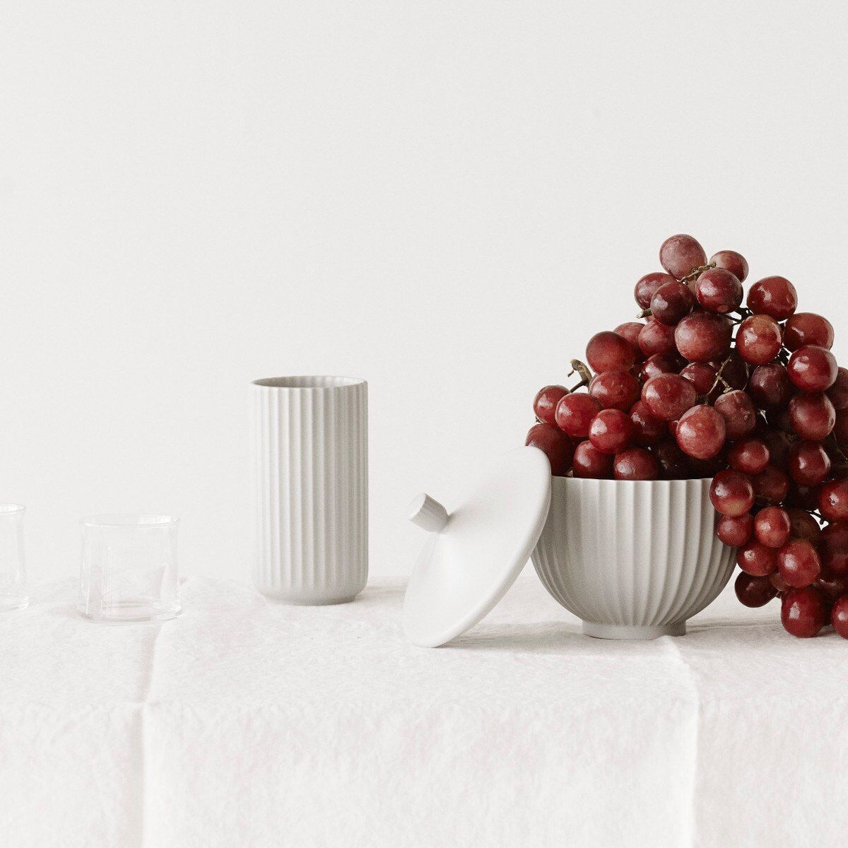 Ein weißer Keramikvase »Lyngby« mit einem gerippten Design, gefüllt mit roten Weintrauben. Auf einer weißen Tischdecke steht sie, umgeben von drei leeren, klaren Gläsern.