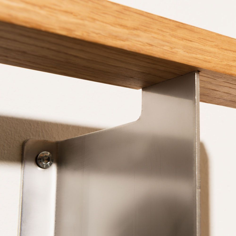 Detailansicht eines Regals namens »b-eck«, das eine Kombination von Holzregalboden und einer stabilen Metallhalterung zeigt. Die Holzplatte ist hellbraun, ergänzt durch eine silberne Metallhalterung an einer weißen Wand.