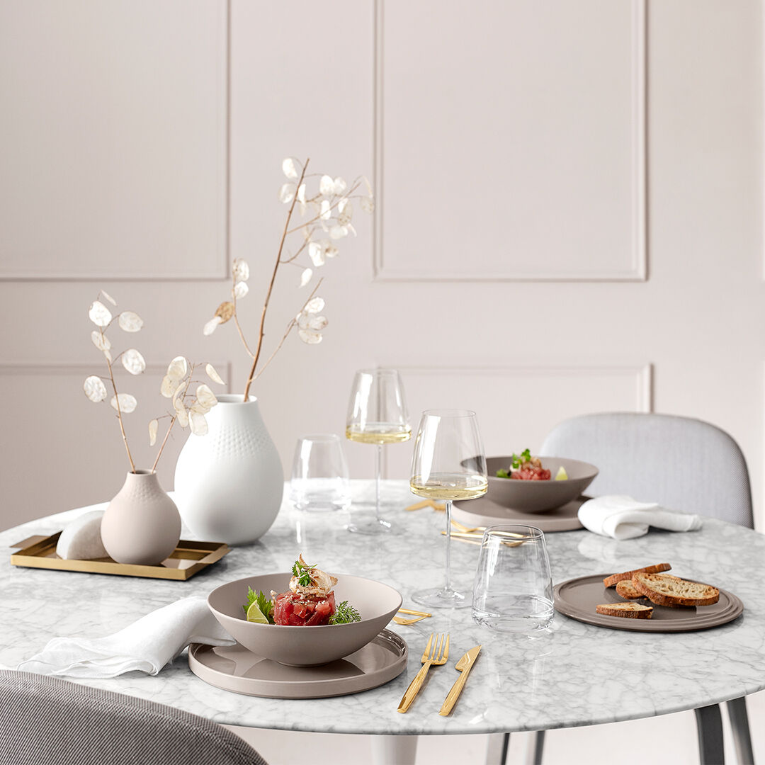 Ein elegantes Geschirr-Set, genannt »La Boule«. Das Set beinhaltet unter anderem Teller, Schalen und Gläser, die auf einem runden Marmortisch arrangiert sind. Die Tischdekoration ist minimalistisch und modern, mit neutralen Farbtönen in Weiß, Beige und Grau sowie goldenen Akzenten. Ein schönes Setting für ein feierliches Essen mit weißen Blumen in einer Vase und elegantem Essgeschirr.