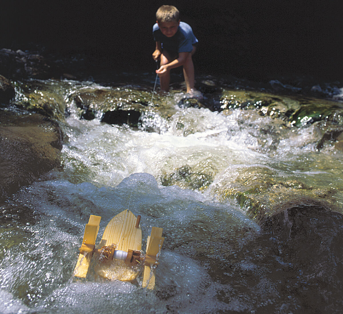 Ein Kind experimentiert draußen an einem fließenden Bach mit einem Bausatz in Form einer Forelle. Das Wasser spritzt und bewegt sich um den gelben Bausatz herum, während das Kind konzentriert zusieht.