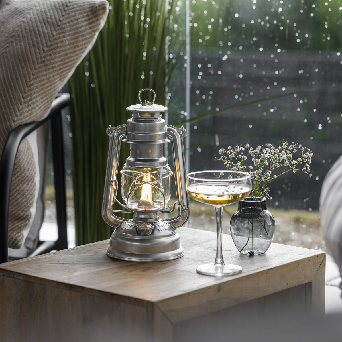LED Laterne »Baby Special 276« steht auf einem Holztisch neben einem Glas mit Getränk und einer kleinen dekorativen Vase. Im Hintergrund ist das Zimmer durch ein Fenster im Regen sichtbar. Die Szene wirkt gemütlich und entspannend.