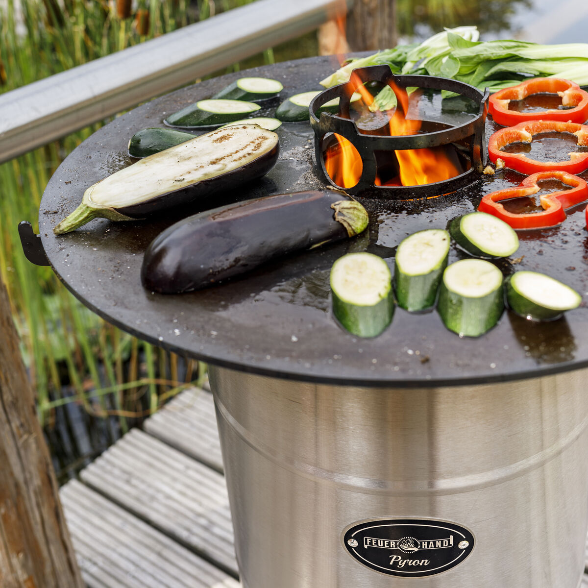 Ein silberner Pyron Grill mit einer schwarzen Grillplatte, auf der verschiedene Gemüsesorten wie Zucchini, Paprika und Auberginen gegrillt werden. Im Hintergrund ist ein natürlicher Teich zu sehen.