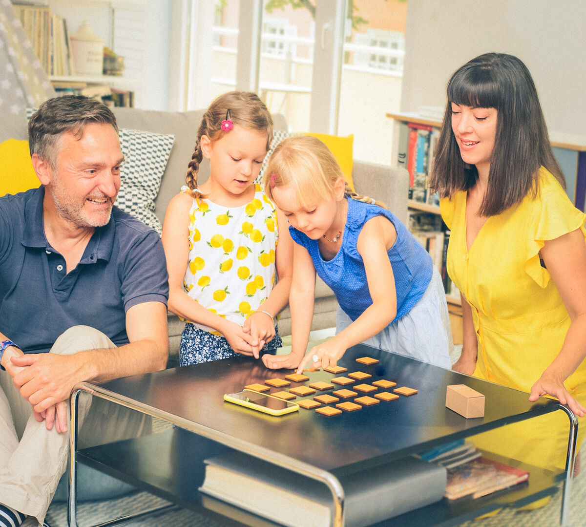 Eine Familie sitzt an einem Tisch und spielt gemeinsam das Spiel 'klang² Spielesammlung »Geigenbauer«'. Zwei Kinder stehen in der Mitte und greifen nach Spielsteinen auf dem Tisch, während die Eltern lächelnd zuschauen. Im Hintergrund sind Regale mit Büchern und hell erleuchtete Fenster zu sehen.