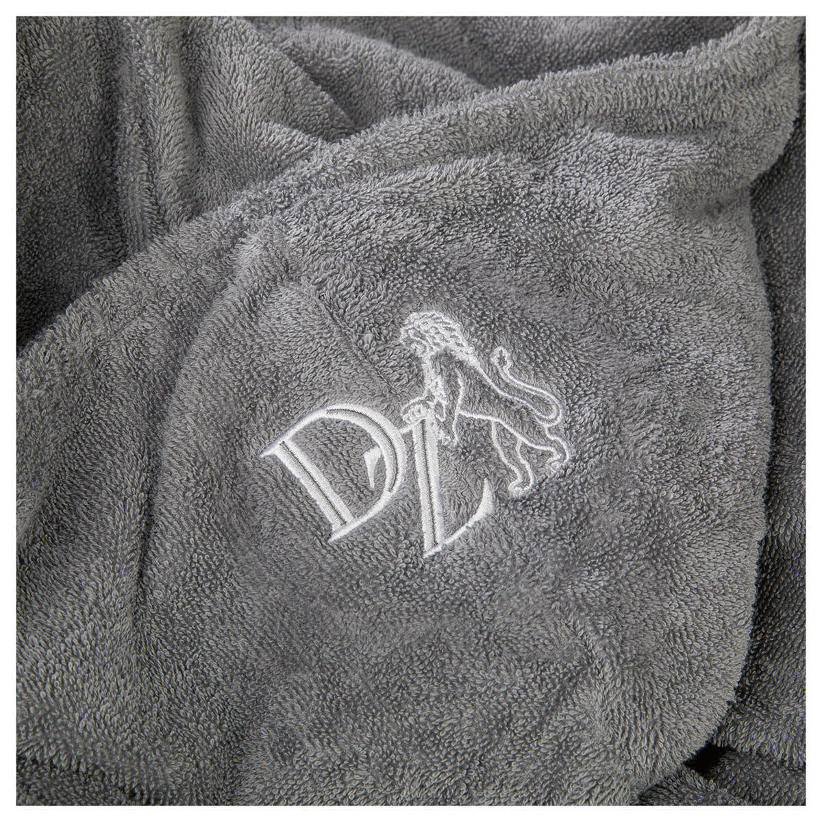 Nahaufnahme eines grauen Bademantels aus Frottee mit einer weißen Stickerei. Die Stickerei zeigt einen Löwen und die Buchstaben 'DL'. Der Bademantel sieht weich und komfortabel aus.