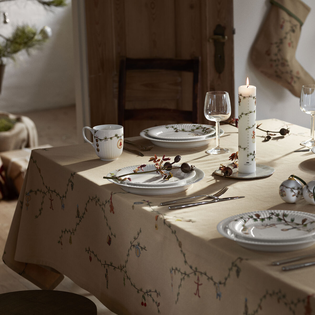 Beigefarbene Tischdecke mit feinem Weihnachtsmotiv auf einem gedeckten Tisch, dekoriert mit Kerzen, festlichem Geschirr, Gläsern und einem Weihnachtskuchen in einem hellen, gemütlichen Esszimmer.