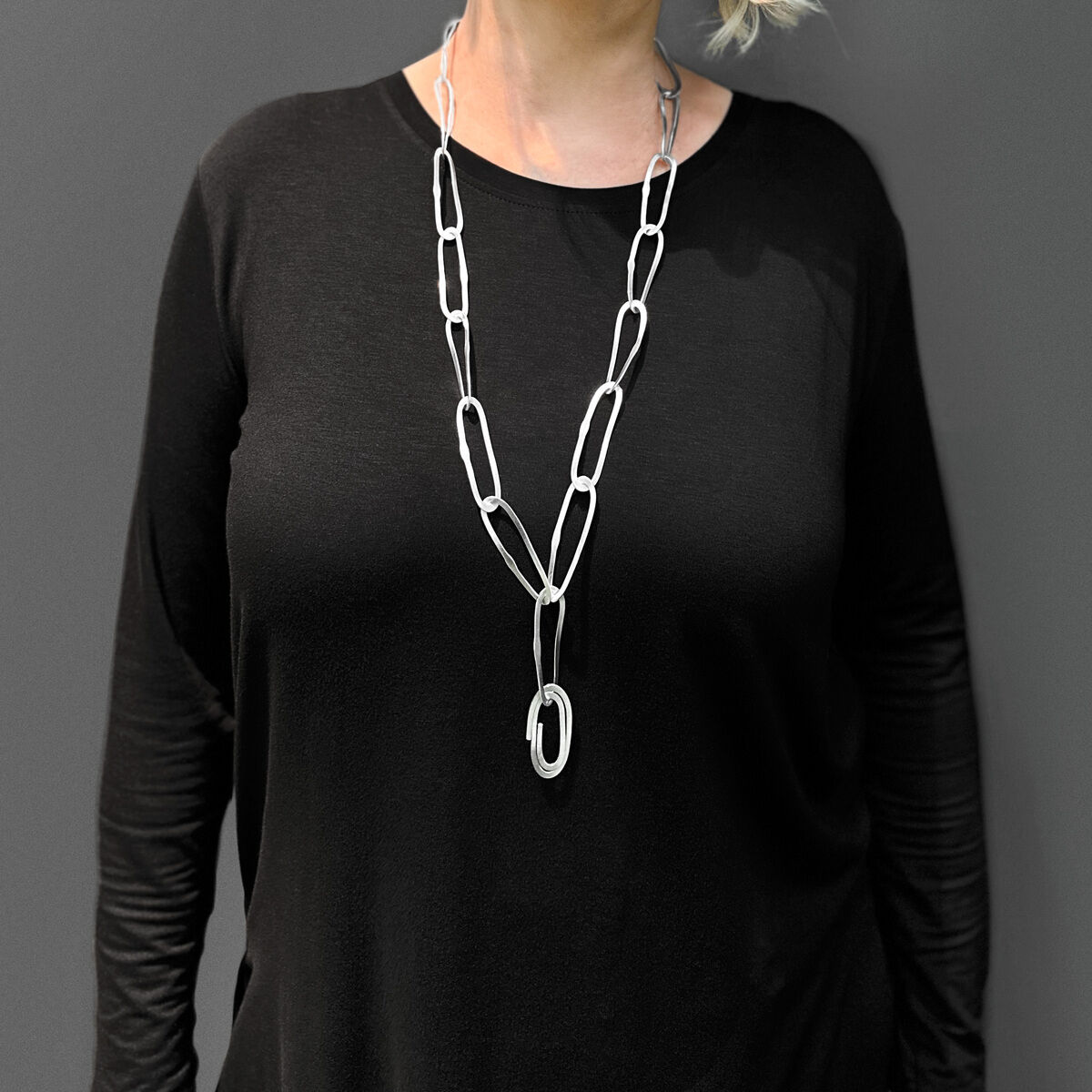Dame mit blonder Frisur trägt eine lange silberne Halskette mit wellenförmigem Anhänger vor einem grauen Hintergrund, gekleidet in einem schwarzen Oberteil.