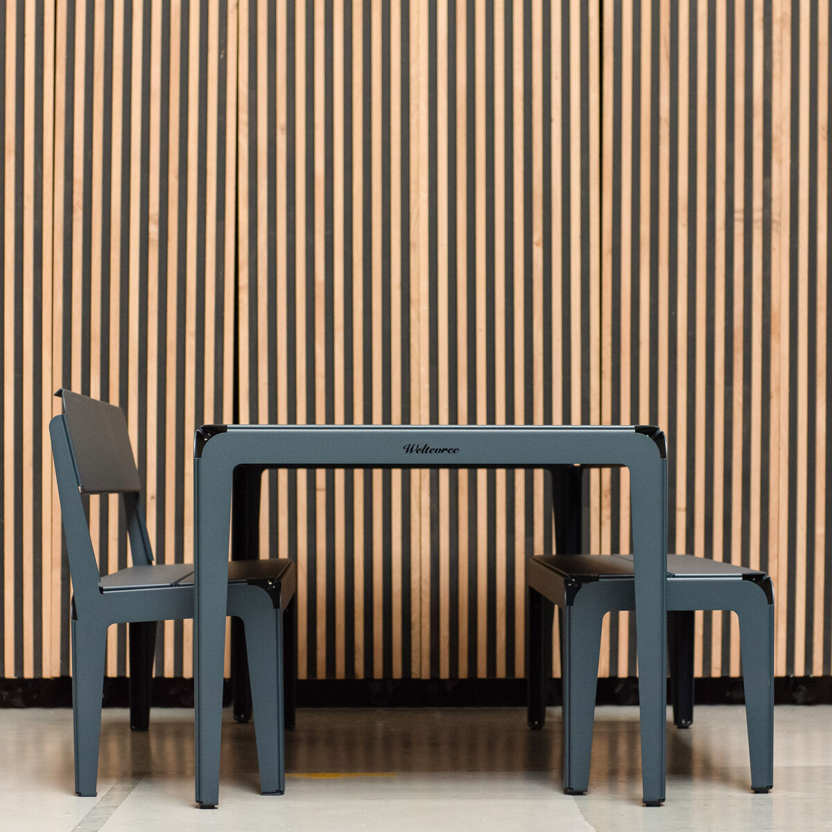 Quadratischer Bended Gartentisch mit zwei passenden Stühlen, vor einem Hintergrund mit vertikalen Holzbalken. Der Tisch hat ein modernes und funktionales Design, geeignet für den Außenbereich.