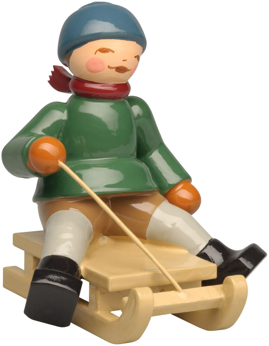 Freigestellte Produktansicht der handbemalten Holzfigur »Winterkinder – Junge auf Schlitten« von Wendt & Kühn. Der Junge trägt eine dunkelblaue Mütze, roten Schal, grünen Mantel, beige Hose, orange Fäustlinge und schwarze Stiefel. Er sitzt auf einem naturholzfarbenen Schlitten und hält eine dünne Zugschnur.