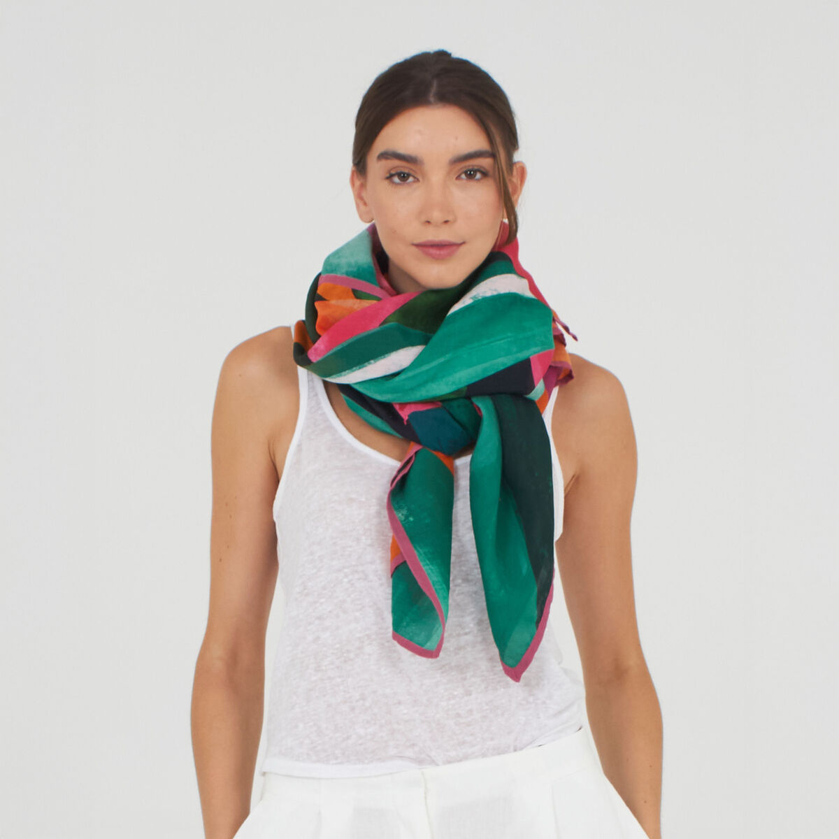 Eine Frau trägt einen auffälligen Baumwoll-Seidenschal "Cassey" von Shalls Barcelona in Orange mit bunten geometrischen Mustern. Der Schal ist voluminös um den Hals drapiert und kombiniert Farbtöne wie Orange, Grün, Türkis und Pink zu weißen Akzenten.