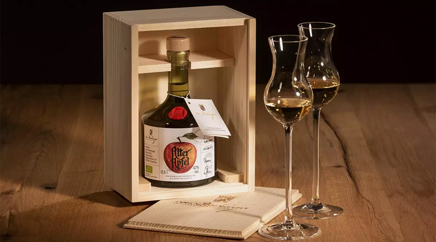 Eine Holzkiste mit einer Flasche „Alter Apfel“-Likör, daneben zwei elegante Gläser, auf einem Holztisch.