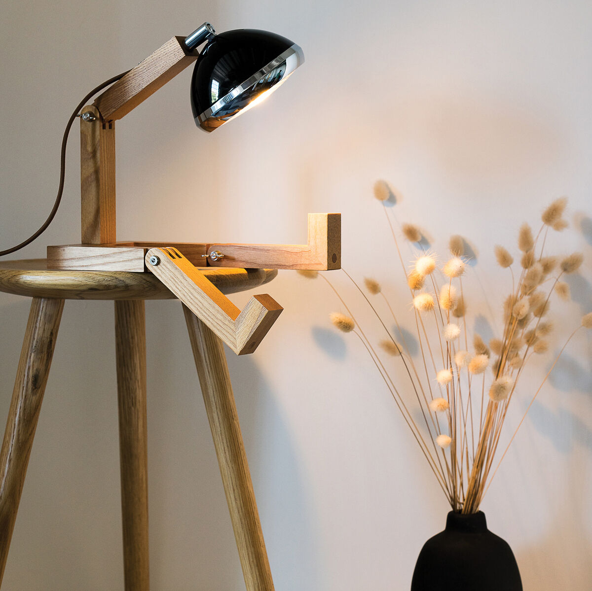 Leuchte »Mr. Wattson« auf einem Holzstuhl neben einer schwarzen Vase mit getrockneten Gräsern. Die Lampe hat ein skandinavisches Design mit verstellbaren Holzarmen und einem Metallkopf.